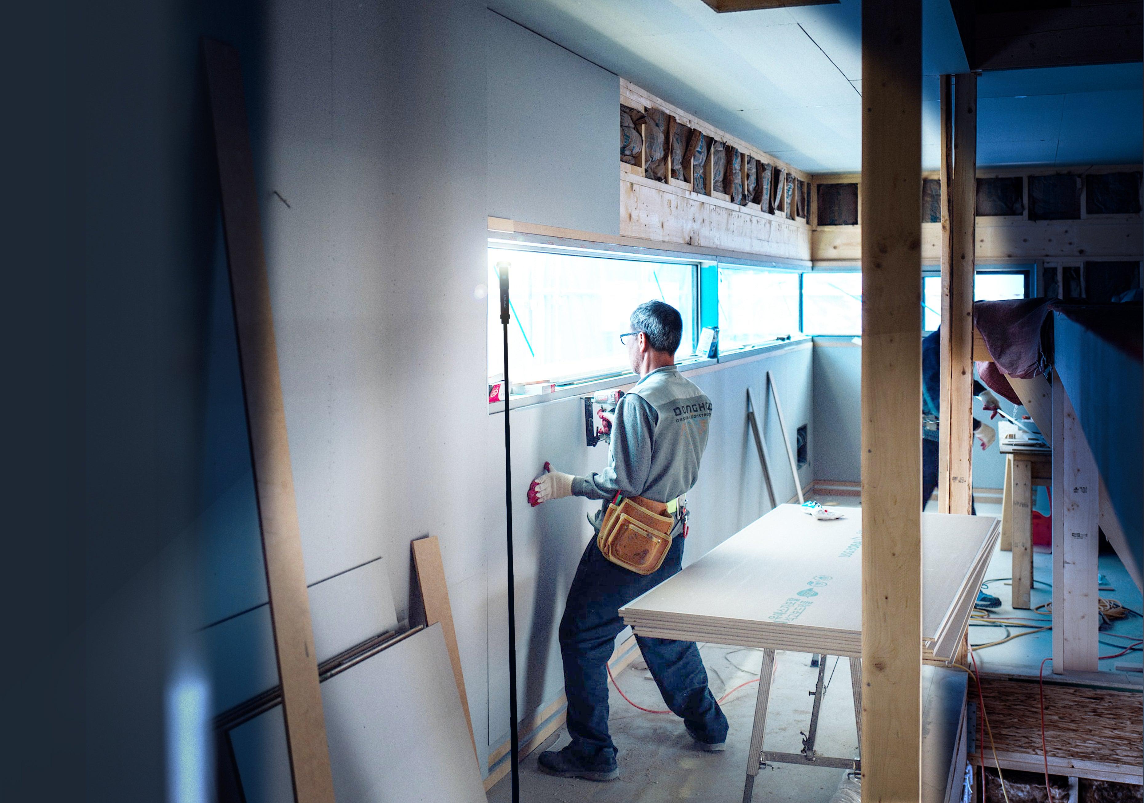 Male construction worker hanging drywall with an air tool. His immediate work area is lit up by a FLi Telescoping Light by STKR Concepts