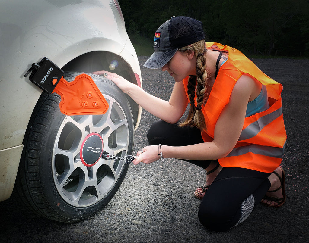 Mujer vistiendo un chaleco naranja de seguridad arrodillada apretando la tuerca del neumático de su vehículo por la noche iluminada por una linterna flexible FLEXIT Auto