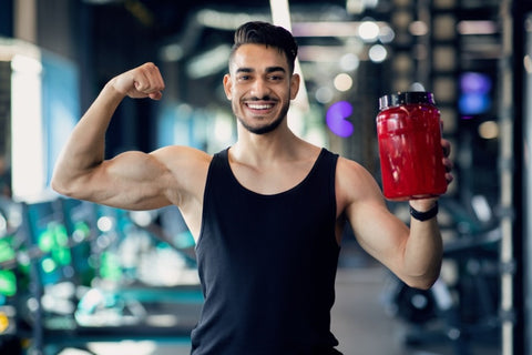 shot of a muscle builder smiling at the camera while holding a red supplement container and flexing his right arm