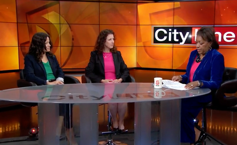 Two women being interviewed by a TV host at a TV news desk