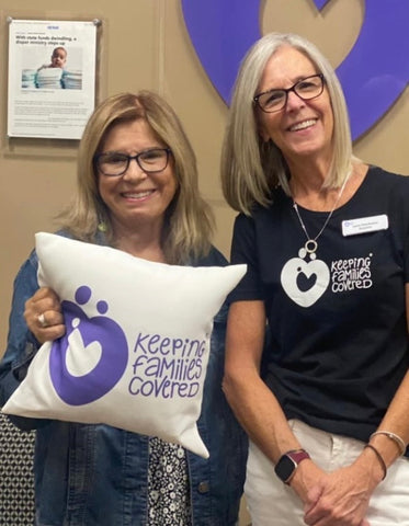 One woman holding Keeping Families Covered pillow and one woman wearing Keeping Families Covered black t-shirt