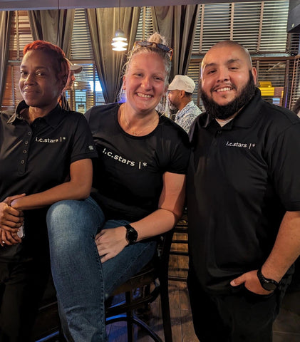 In a bar together, one woman wearing i.c.stars black polo shirt; one woman wearing black i.c.stars black t-shirt; one man wearing black i.c.stars black polo shirt