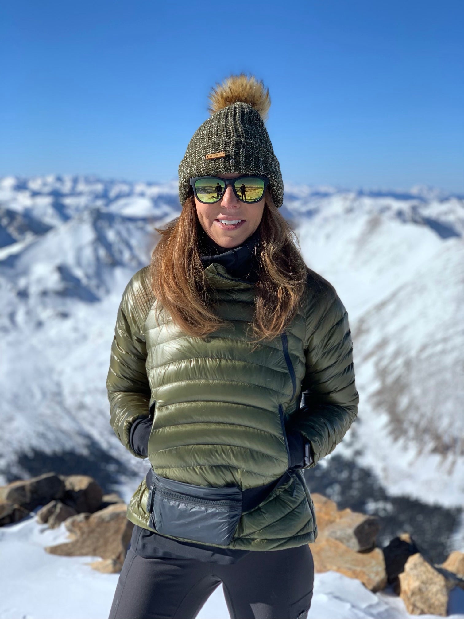 Smiling woman standing at top of snowcapped mountain wearing a fanny pack