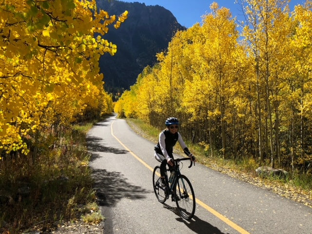 Woman biking on paved path with bright yellow trees and mountain behind her