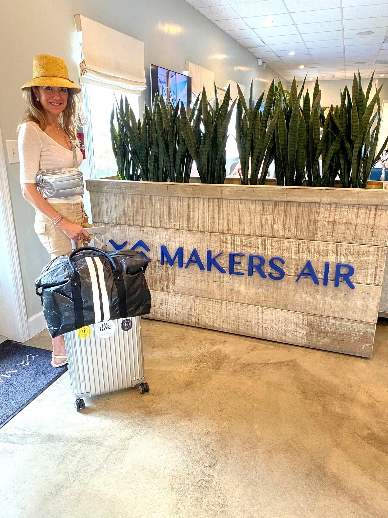 Woman in front of Makers Air sign with weekender bag on top of silver suitcase