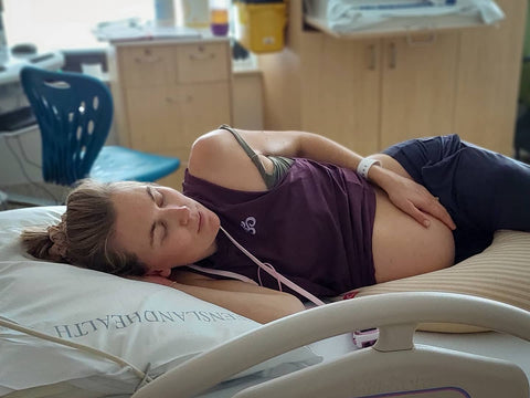Woman lays on hospital bed calmly birthing with tens machine on lanyard around her neck and her hand gently resting on her belly