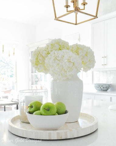 Display in kitchen with white cabinets and counter tops – there is a cream/white tray on the counter with 3 White cluster of flowers in a white vase, green apples in a white fruit bowl and a candle in a glass container. Interior design blog about our tips on bringing your kitchen to life without spending too much money. Written by vintage-etc in Cape Town. We make custom made & bespoke tables, benches, free-standing kitchen islands, desks, media units & wardrobes in our Cape Town & Johannesburg workshops – using Oak, Oregon, Ash, Beech, Birch Ply & Meranti. We also sell imported furniture, provide design consulting services & make soft furnishings e.g. sofas & upholstered chairs in linen, velvet & stain resistant fabric.
