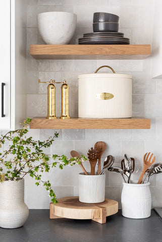 White textured tiled kitchen wall with Solid Oak floating shelves displaying bowls and a bread bin. Underneath the floating shelves on the black counter top are wooden spoons and metal utensils in a white pot and a pot plant in a ceramic pot. Interior design blog about our tips on bringing your kitchen to life without spending too much money. Written by vintage-etc in Cape Town. We make custom made & bespoke tables, benches, free-standing kitchen islands, desks, media units & wardrobes in our Cape Town & Johannesburg workshops – using Oak, Oregon, Ash, Beech, Birch Ply & Meranti. We also sell imported furniture, provide design consulting services & make soft furnishings e.g. sofas & upholstered chairs in linen, velvet & stain resistant fabric.