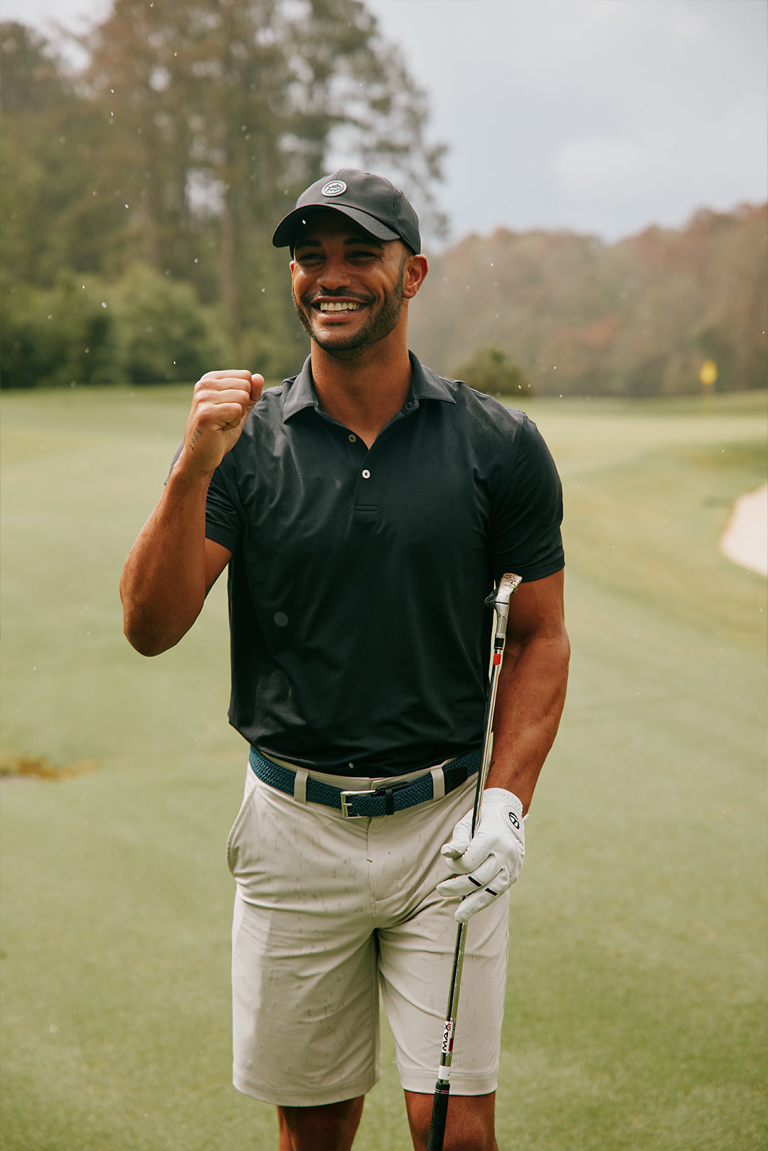 Man in black polo shirt and khaki shorts on a golf course celebrating his shot.