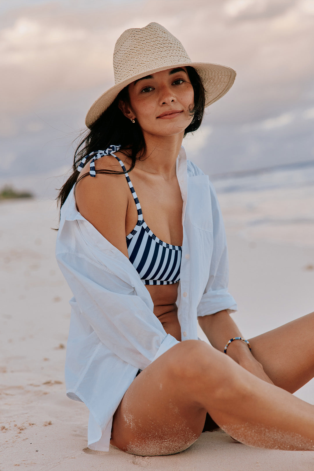 Woman on the beach in a striped bikini and white coverup wearing a hat.