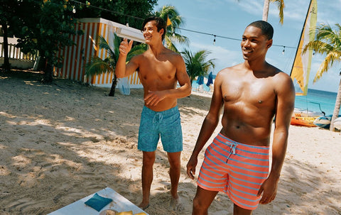 Two men playing cornhole on the beach in swim trunks.