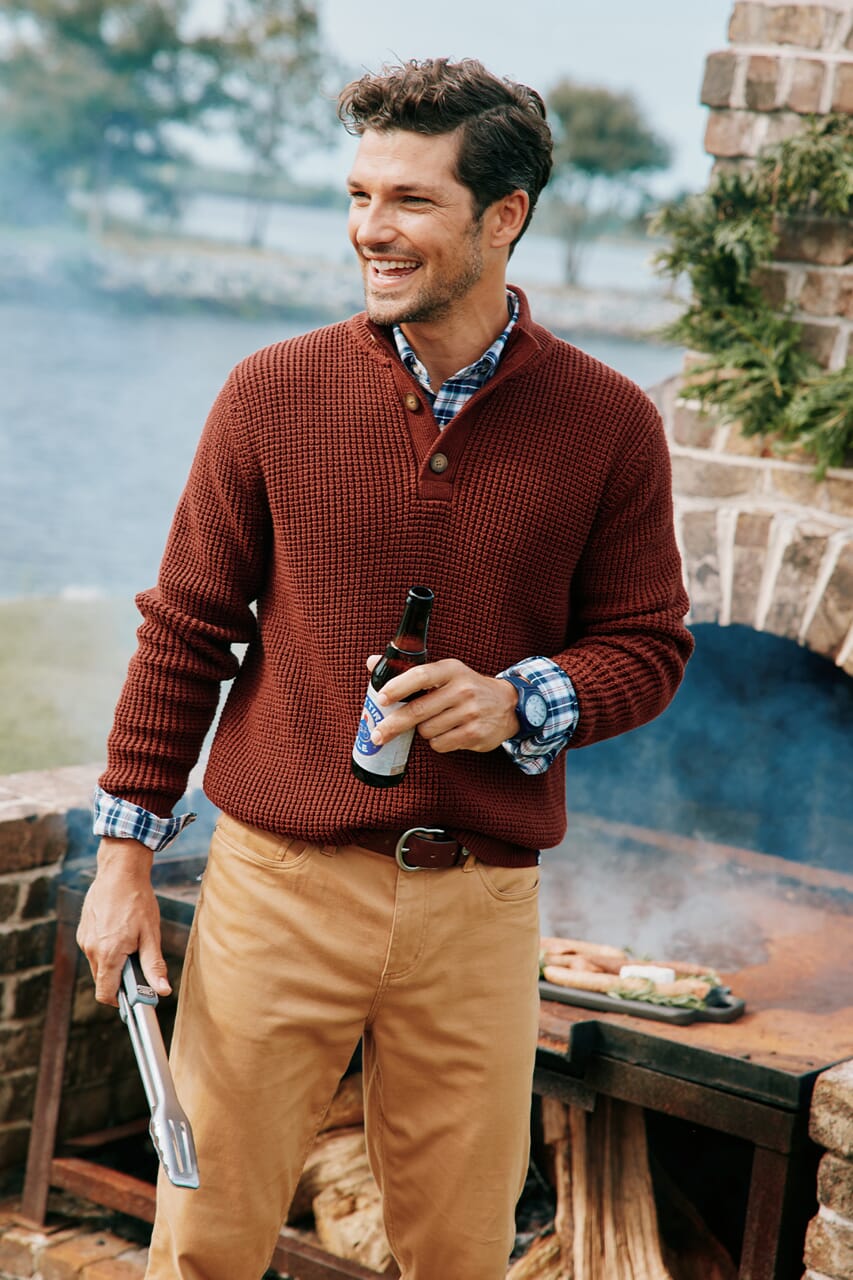 Man in khaki pants at a bonfire in the fall with a red pullover quarter zip and plaid button up shirt.