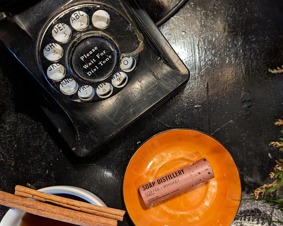 image of coffee + whiskey lip balm in a brown paperboard tube, lying on a small orange plate next to a black dial up phone and a mug of tea