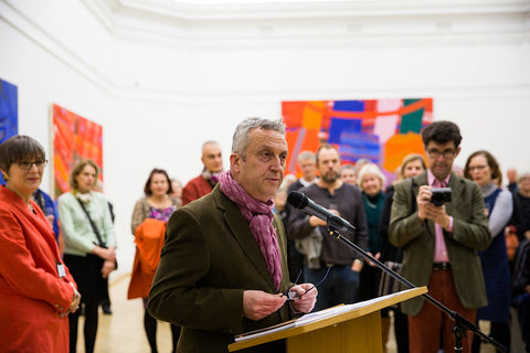 Stewart Geddes speaks into a microphone at the RWA, people stand behind him, with bright abstract paintings on the wall in the background