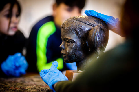 An image of a hand resting on sculpture of a head, in the background two children watch