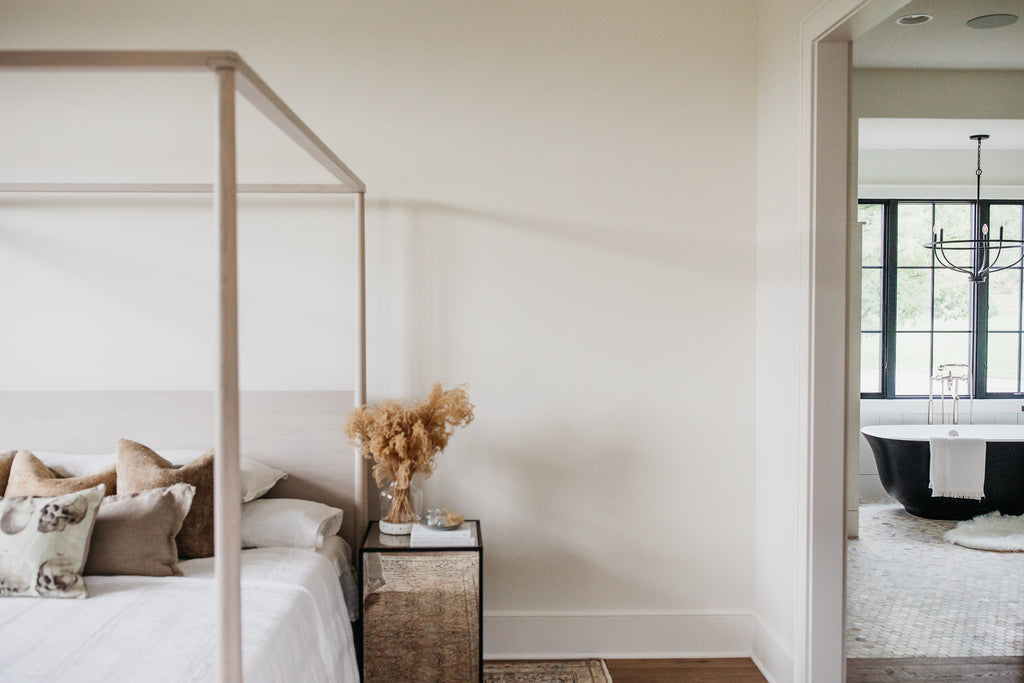 Master bedroom with whitewashed wood canopy bed, pom pom linen bedding, iron cushioned chairs, and vintage persian rug.