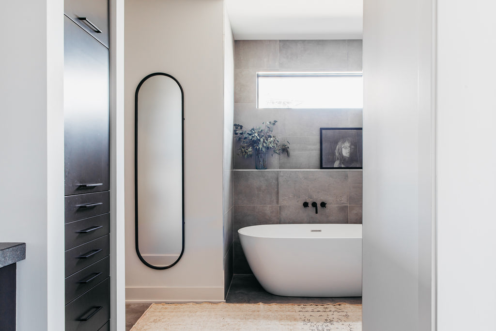 Salle de bain principale de type spa avec miroir en fer de forme ovale, tapis sable et baignoire blanche.