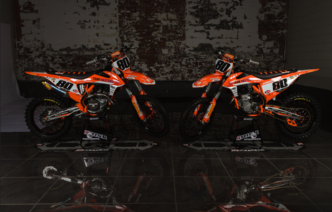 Professional motocross/supercross racer Kevin moranz race bikes being picture in a photo shoot in a dark brick walled room with his bikes sitting on the A.T.S. stand and factory pit mat