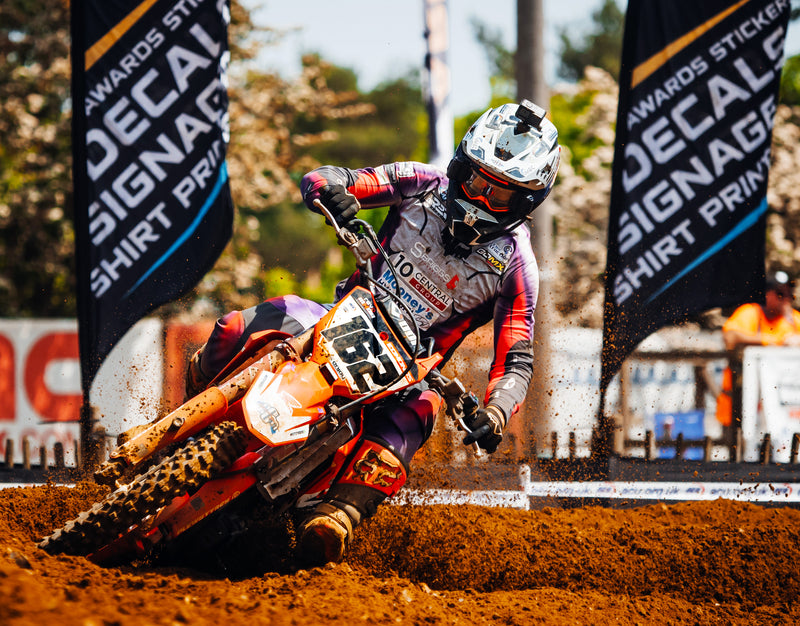 motocross racer leaning hard as he navigates a soft dirt turn. He's coming straight at camera so we can only see the front plews tire. Two huge race promotional flags in the background.