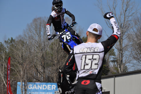 Motocross rider offering his peer encouragement from the side of the track in the form of a fist pump while his friend flies through the air in style on his yamaha dirt bike.