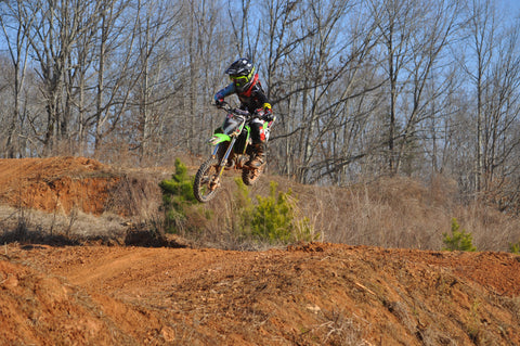 RISK Racing athlete training on a motocross. The rider is wearing the necessary motocross gear, which includes risk racing pants, jersey, gloves, and goggles. The riders gear is is red teal, yellow and black the goggles are green, and the dirt bike they are riding is also green. The motocross rider is mid air after leaving the take-off of the jump.