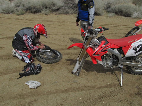 Trial riders experiencing a flat front tire on the trail. the two riders have removed the front wheel from the bike, as well as the punctured tube and installing a new tube so they can repair the flat tire and cintinue their ride.