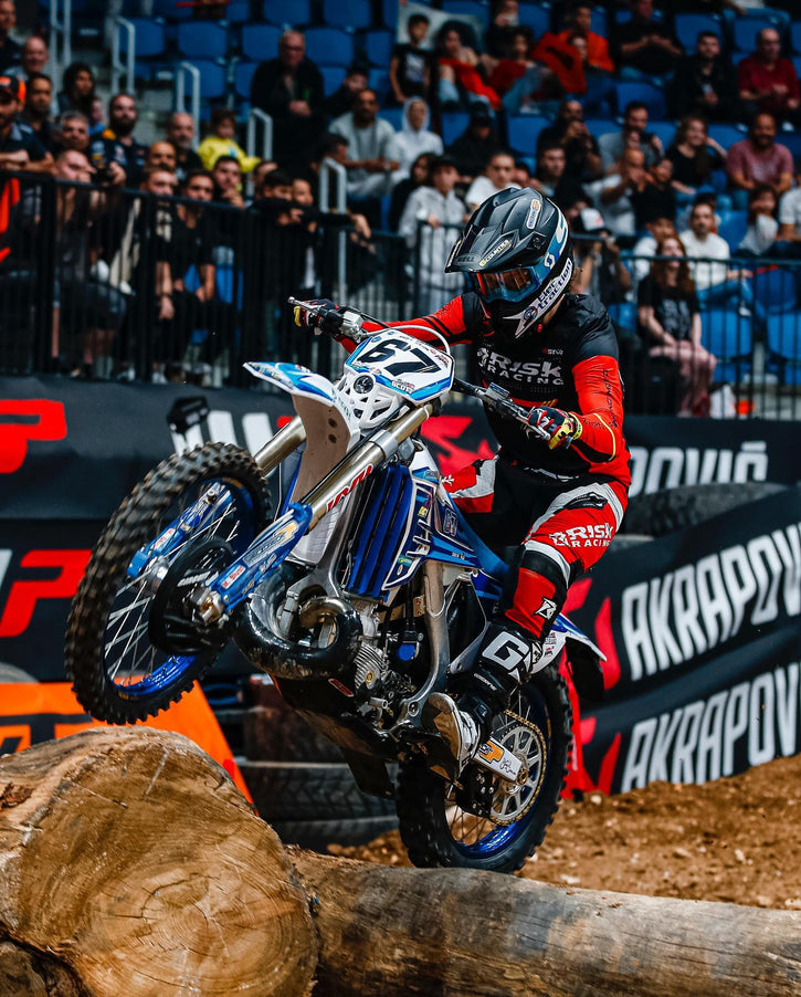 Endurocross Racer che lancia su alcuni tronchi in pista. Indossa un attrezzatura da corsa per il rischio e la sua bici è dotata di pneumatici plews. I fan dello stadio in sottofondo.