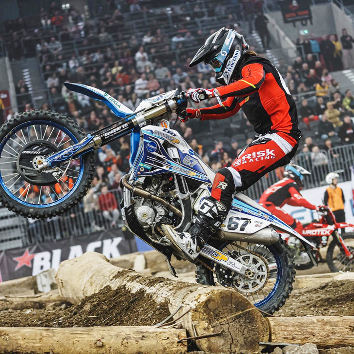 Enduro Racer mid hop over a huge log. He's wearing Risk Racing riding gear and running Plews Tyres. Stadium of fans in the background.