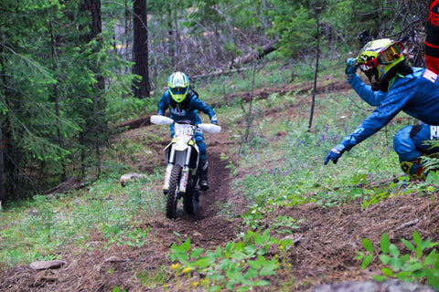 Learn to ride a dirt bike confidently. Riding a white dirt bike in a lush green forest up a steep hill with encouragement from their peers to keep going. 