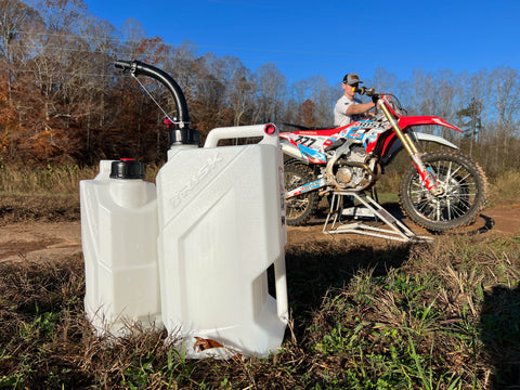 Jarras utilitarias fotografiadas junto a una pista de motocross frente a motos de cross sentadas en el césped en un día de cielo azul. Las jarras utilitarias son de color blanco transparente con mangueras negras en la parte superior.