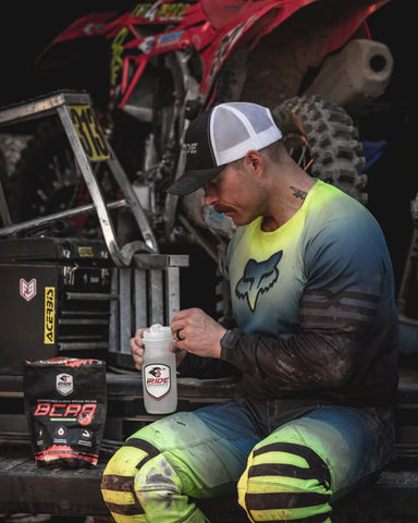 Un athlète de motocross remplissant sa bouteille d'eau avec des suppléments iRide de BCAA tout en étant assis sur le pare-chocs de sa moto van. Vélo de motocross dans la camionnette.