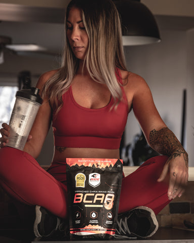 Mujer sentada con las piernas cruzadas bebiendo BCAA después del entrenamiento en una botella agitadora y vistiendo un traje de entrenamiento granate.