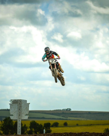 Piloto de RISK Racing en su ktm 250sxf volando por el aire después de alcanzar el despegue de una gran característica en una pista de motocross. No hay ninguna parte de la pista que se muestre debajo del ciclista, solo la ladera al fondo. El piloto lleva equipo de motocross ventilado v2 de Risk Racing.