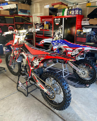 two dirt bikes, one sitting on a risk racing a.t.s. stand the other resting on a risk racing ride-on lift. The garage has a great moto set up with race numbers on the wall $ organized tool drawers,