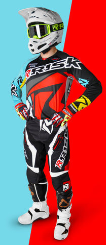 person standing in risk racings red/teal ventilate v2 mix-n-match series gear with the v3 green goggles in a white helmet and white boots. The person is standing with their hands on their hips.