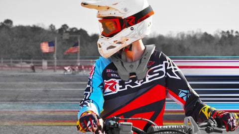 Motocross rider sitting on bike at a standstill with hands on the handle bars wearing black, red, and teal risk racing motocross jersey, gray neck brace, white helmet and red goggles, the most important pieces to this kit is the neck brace and helmet.