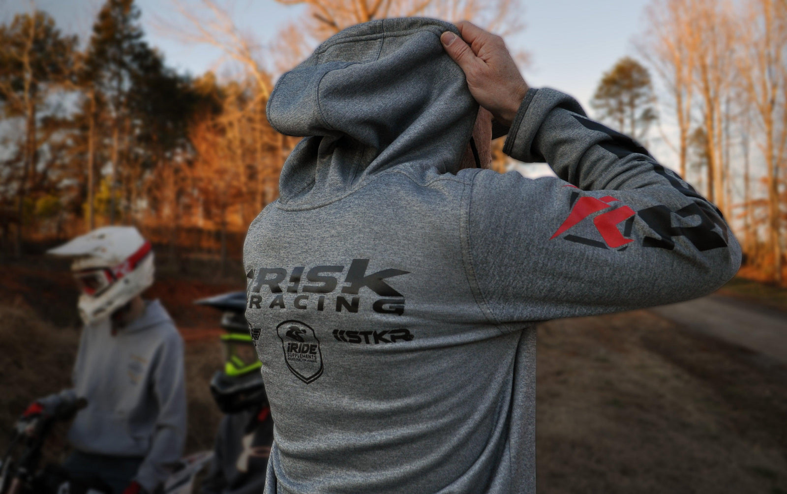 Parte trasera de un hombre que lleva una chaqueta con cremallera Risk Racing y se pone la capucha en la cabeza. Dos jóvenes pilotos de MX y una escena de tierra/árbol se difuminan en el fondo.