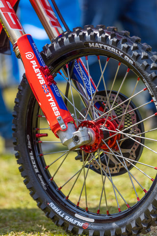 Pictured is the front tire of a motocross race bike, This front tire is the Matterly GP intermediate terrain tire form PLEWS Tires. The wheel the tire is mounted to has anodized red hubs, anodized red spoke nipples on black wheels, in the background is grass and another race teams pit set up.