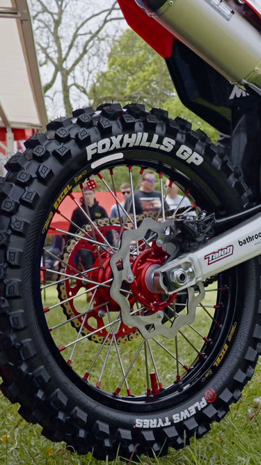 Primo piano della gomma posteriore e del braccio di swing di una bici da motocross in un ambiente di erba esterna. Le lettere di Whitewall di Foxhills GP e Plews hanno mostrato in modo prominente le gomme.