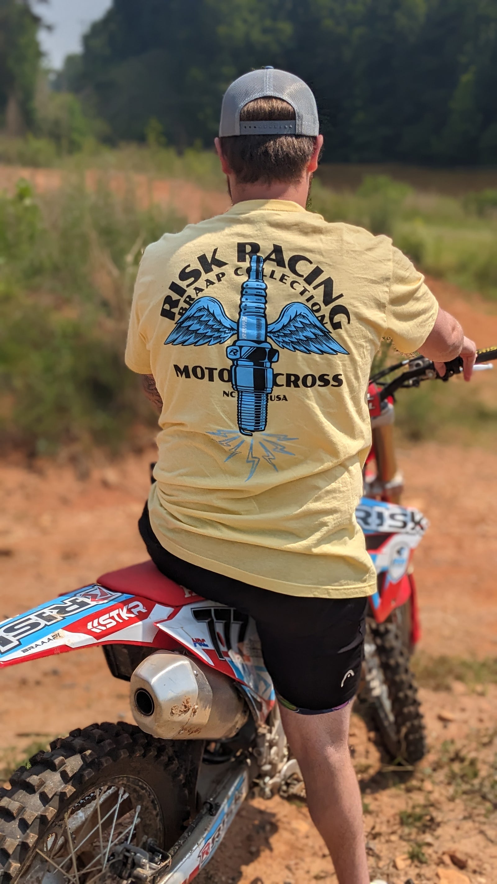 Hombre sentado en una moto de cross de espaldas a nosotros mostrando su camiseta amarilla de Risk Racing con alas de chispa.