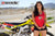 Le modèle de moto de mars de Risk Racing, Amber Juliana, portant un débardeur rouge Risky moto chick daisy tirant au bas de celui-ci avec les deux mains tout en posant devant un vélo de motocross - gros plan - piste MX clôturée blanche en arrière-plan
