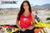 La modelo de moto de marzo de Risk Racing, Amber Juliana, con una camiseta sin mangas roja Risky moto polluelo margarita tirando de la parte inferior con ambas manos mientras está de pie frente a una bicicleta de motocross - primer plano - cerca blanca de la pista MX en el fondo