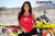 La modelo de moto de marzo de Risk Racing, Amber Juliana, vestida con una camiseta sin mangas roja Risky moto polluelo daisy y pantalones cortos negros con ambas manos cerca de la cintura mientras posa frente a una bicicleta de motocross - primer plano - cerca blanca de la pista MX en el fondo