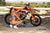 Risk Racing's May Moto Model Natalie Nicole wearing a 2 piece bikini kneeling sideways in front of a motocross bike that's sitting on an ATS Stand and Factory Pit mat by Risk Racing. Her left hand on the seat and right resting on her knee. Turning her buns towards camera - wide shot - white fenced off MX track in background
