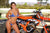 Risk Racing's May Moto Model Natalie Nicole wearing a 2 piece bikini posing in front of a motocross bike that's sitting on an ATS Stand by Risk Racing. Her left hand on the seat and right is slightly pulling up on her bikini bottom strap. - medium close shot - white fenced off MX track in background