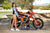 Risk Racing's May Moto Model Natalie Nicole wearing a Teal/Black VENTilate MX Jersey and blue bikini bottoms standing sideways with her right hip to camera in front of a motocross bike resting her left hand on the seat - wide shot - white fenced off MX track in background