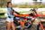 Risk Racing's May Moto Model Natalie Nicole wearing a Teal/Black VENTilate MX Jersey and blue bikini bottoms standing in front of a motocross bike resting her left hand on the seat and her right arm across her torso - medium/wide shot - white fenced off MX track in background