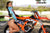 Risk Racing's May Moto Model Natalie Nicole wearing a Teal/Black VENTilate MX Jersey and blue bikini bottoms standing sideways in front of a motocross bike resting her left hand on the seat and her right arm across her mid drift. Her right foot and knee lifted across in front of the bike. - medium/wide shot - white fenced off MX track in background