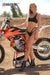 June Moto Model Rochelle Roche wearing a 2 piece black bikini in front of an orange dirt bike with her right hand on the seat. Bike is sitting on an Adjustable Top Stand by Risk Racing. MX track in the background.