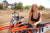 Risk Racing's June Moto Model Rochelle Roche wearing a 2 piece black bikini leaning over an orange dirt bike resting her forearms on the seat. MX track in the background.
